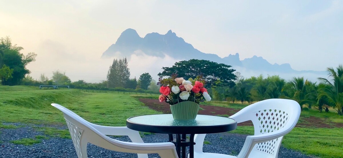 A small round table is set with a decorative vase of flowers, accompanied by two white chairs. In the background, majestic mountains are partially shrouded in mist, creating a serene landscape that reflects the natural beauty of the surrounding area.