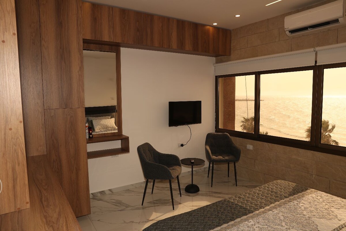A serene studio space features large windows with views of the sea. Two stylish chairs are positioned beside a small table, creating a cozy seating area. A mounted television is located on the wall, and warm wooden accents enhance the design.