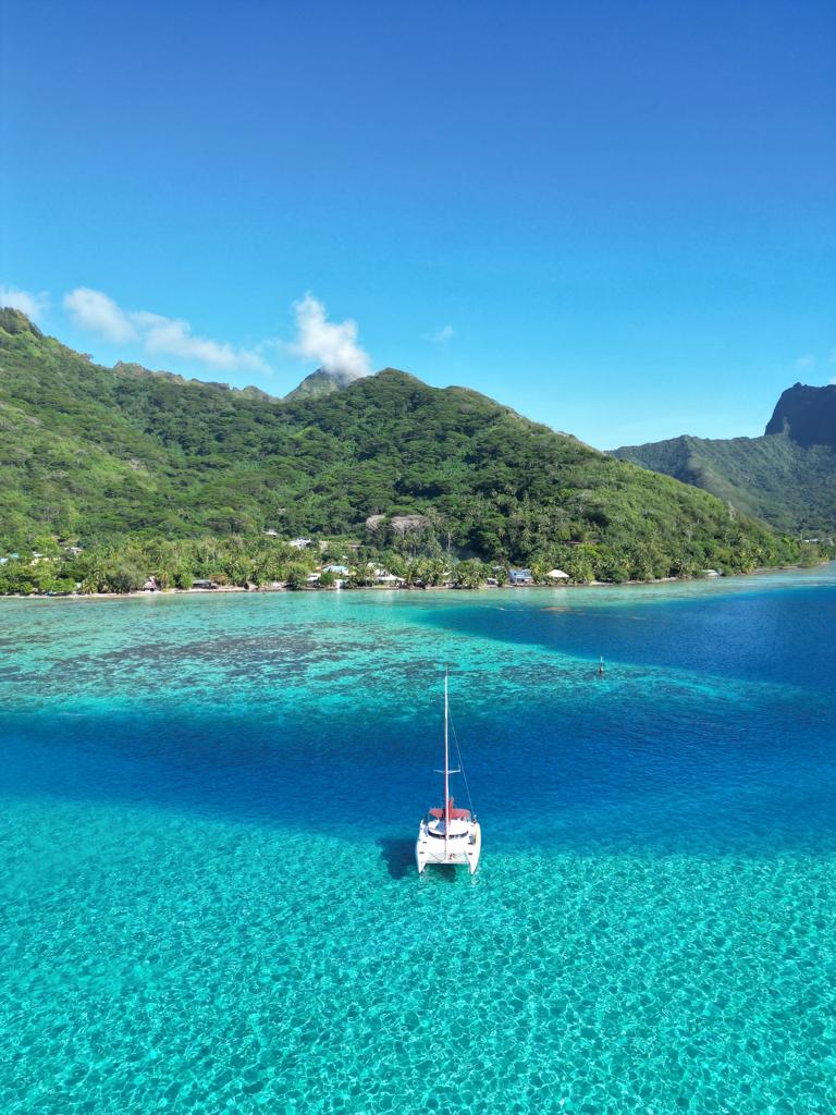 A tranquil scene of clear turquoise waters reflects the surrounding lush green mountains. A sailboat is anchored gracefully in the lagoon, with the shoreline dotted by lush tropical vegetation and distant accommodations enhancing the picturesque setting.