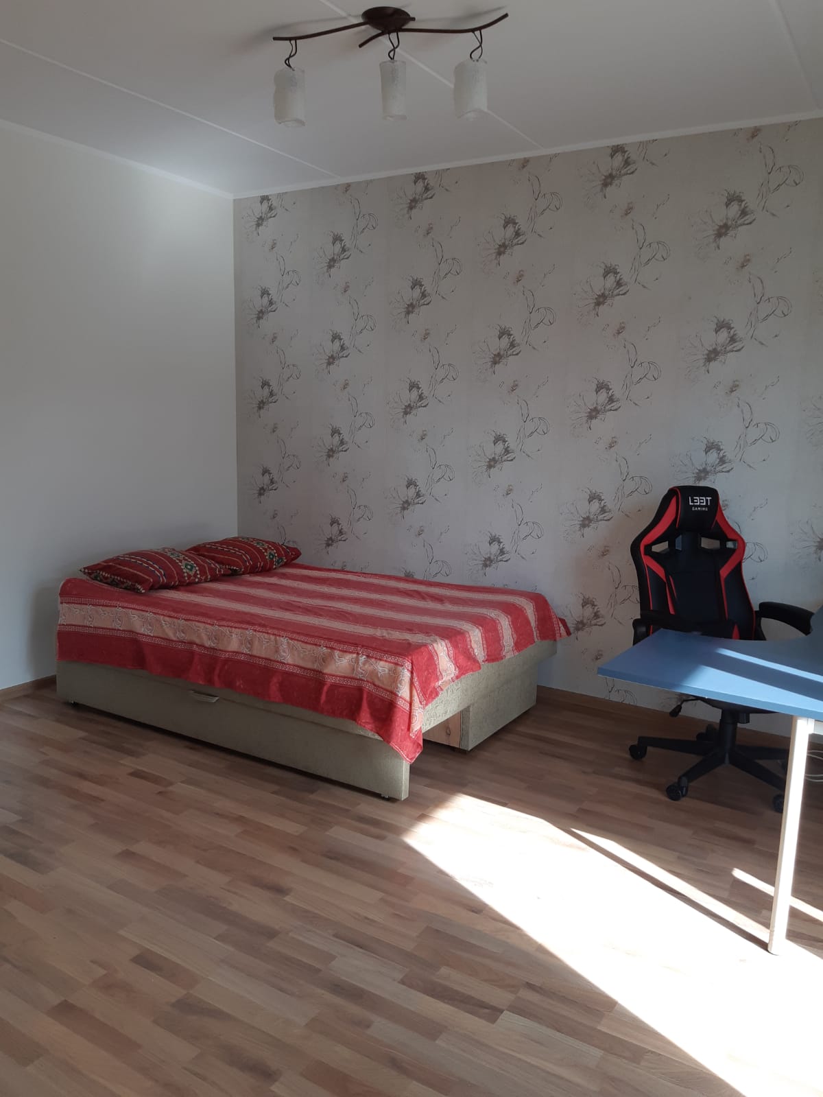 A bedroom is presented with wooden flooring and a feature wall adorned with a subtle floral pattern. A spacious bed, dressed in striped linens, is complemented by two decorative pillows. A desk and an office chair are positioned nearby, with sunlight illuminating the space.
