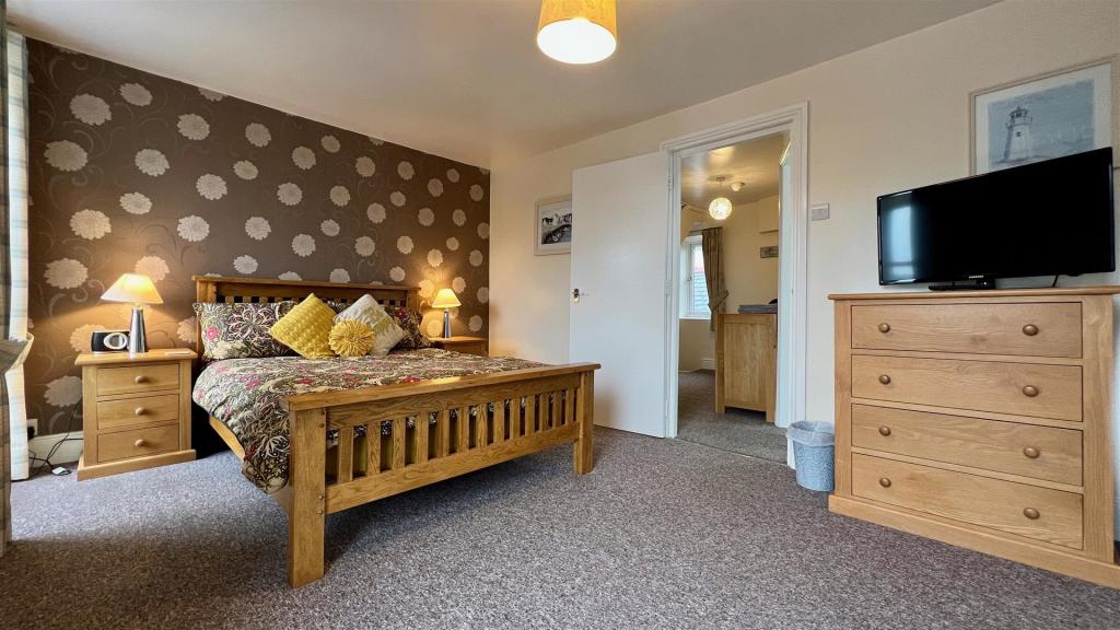 A spacious bedroom features wooden furniture, including a bed with a decorative quilt and cushions. Two bedside lamps provide soft lighting. A television is positioned on a wooden dresser, and a doorway leads to another room. Light filters through a window adorned with curtains.