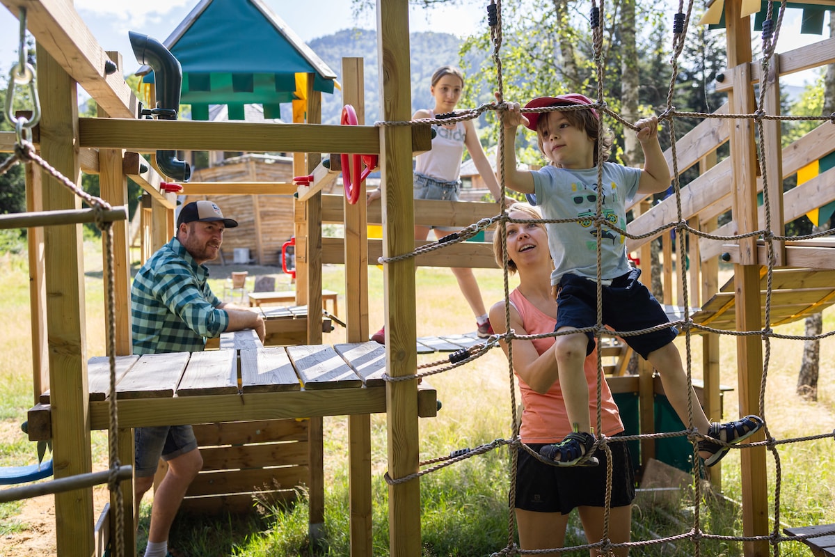 A wooden playground structure is featured, with various climbing and play areas. Children are engaged in play while an adult supervises nearby. The surrounding landscape includes grass and trees, providing a natural setting for outdoor activities.
