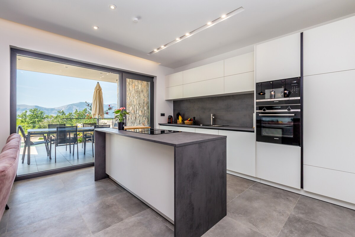 A modern kitchen showcases sleek white cabinetry and a central island with a dark countertop. Large glass sliding doors provide views of the outdoor dining area and surrounding landscape, with natural light illuminating the space. Appliances are integrated into the minimalist design.