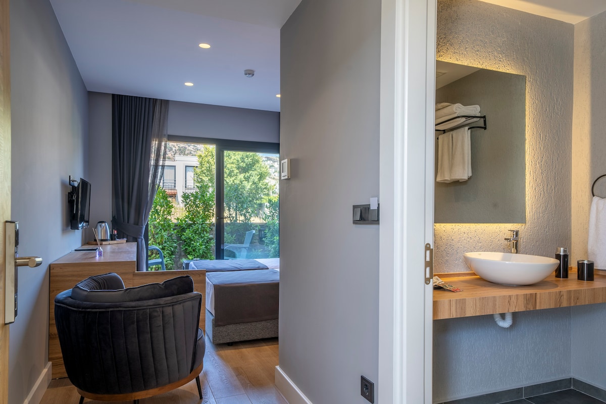 A modern room features a comfortable seating area with a dark armchair and a wooden dresser. A large glass door opens to a private outdoor space filled with greenery. A stylish bathroom area is visible, showcasing a round sink and neatly arranged towels.