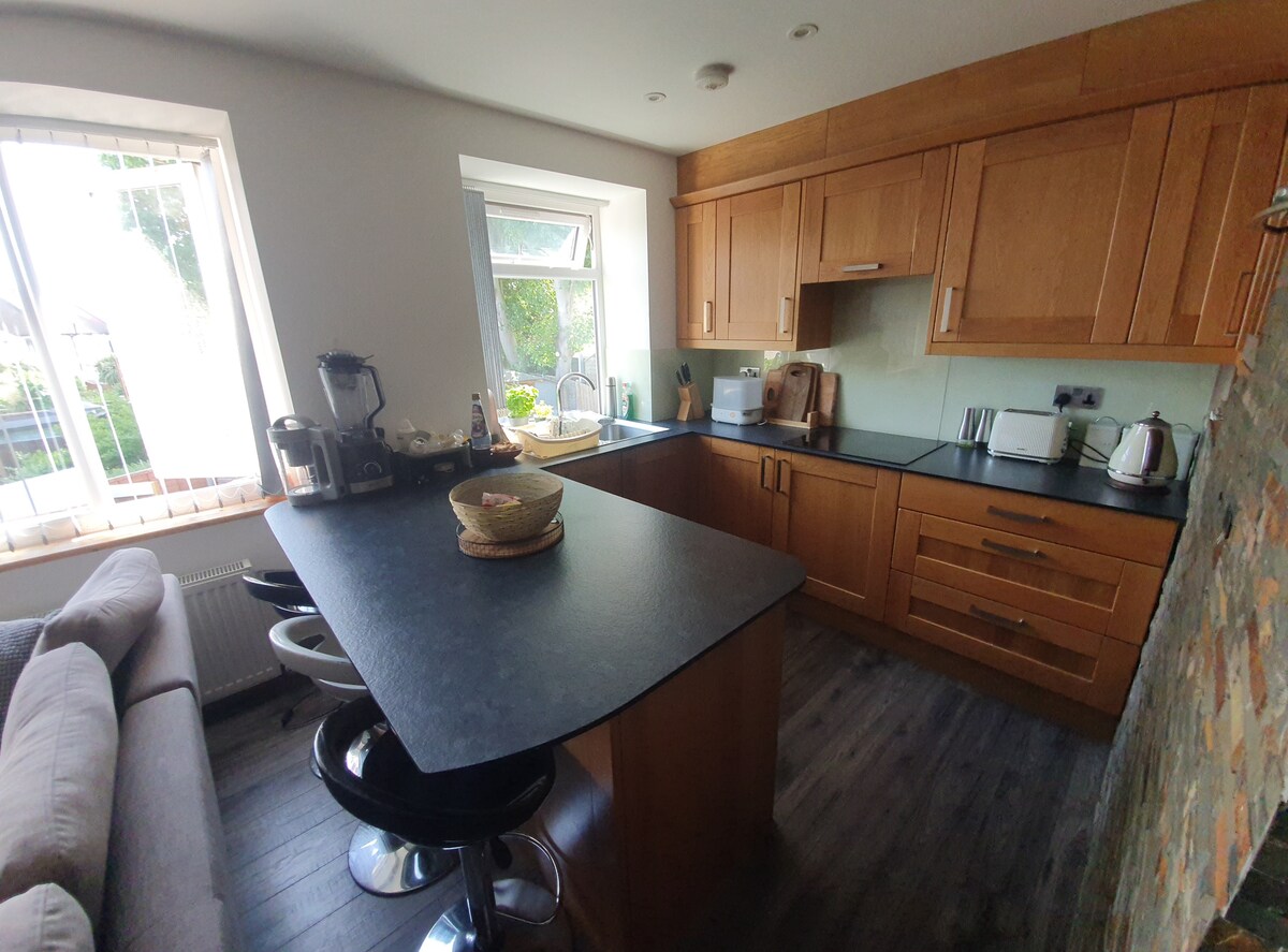 A modern kitchen is featured, equipped with wooden cabinetry and a dark countertop. Bar stools are positioned at a small island, and appliances are visible along with natural light filtering through the windows, highlighting the inviting work area.
