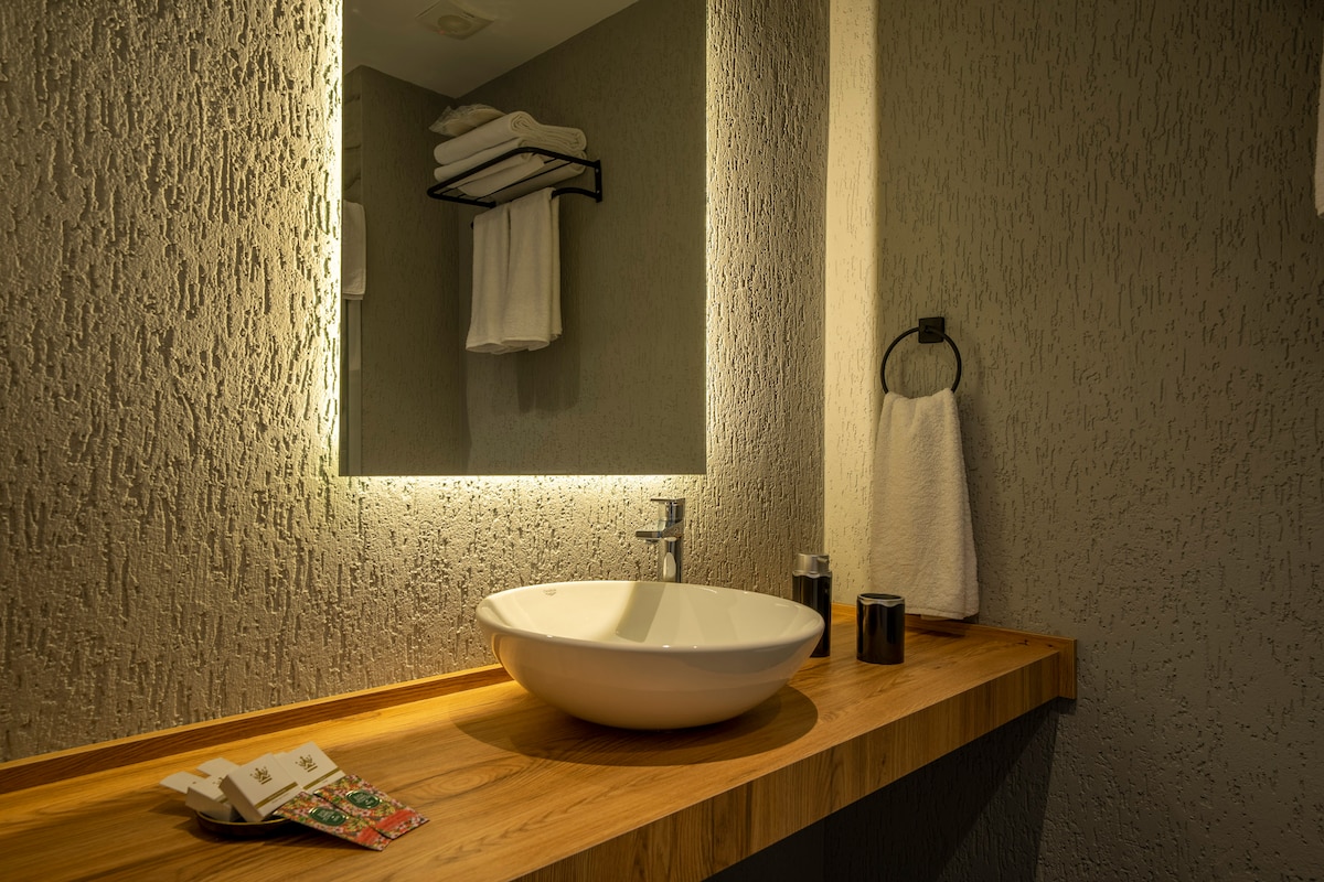 A modern bathroom features a circular sink set on a wooden countertop. A hand towel hangs nearby, and neatly stacked towels are arranged on a black rack above. Soft lighting is provided by a mirror with built-in illumination, enhancing the textured wall.