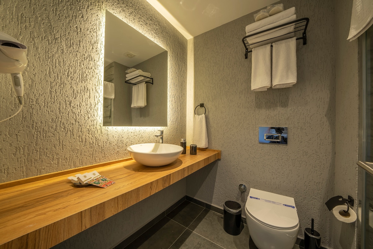A modern bathroom features a spacious wooden countertop with a vessel sink and a wall-mounted mirror illuminated by soft lighting. Fresh towels are neatly stacked on an overhead rack, while a hairdryer is mounted nearby. The floor is tiled in a dark tone, complementing the contemporary design.