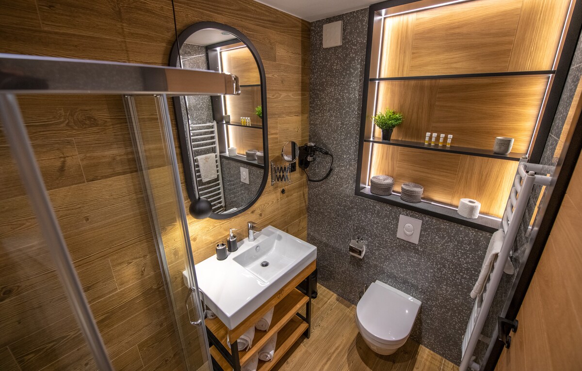 The modern bathroom features sleek wooden accents and textured wall finishes. A stylish vanity is paired with a round mirror, and a walk-in shower is clearly visible. Ambient lighting highlights neatly arranged toiletries on open shelving, enhancing the elegant design.