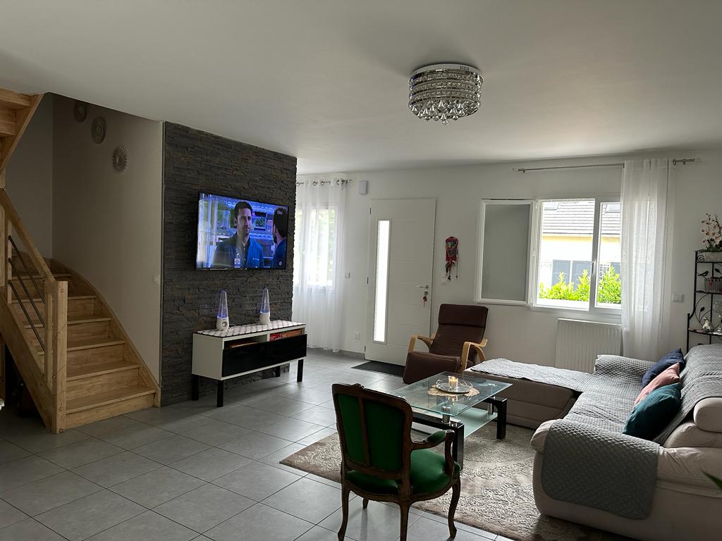 A spacious living area is highlighted by a modern TV mounted on a textured wall. A comfortable sofa and a classic armchair provide ample seating. Natural light enters through the large windows, and the room features a tasteful arrangement of decor, including plants and candles.