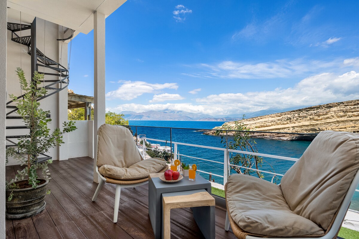 A spacious balcony features comfortable seating arrangements with two soft chairs and a small table. Scenic views of the turquoise sea, distant mountains, and the coastline can be seen. A spiral staircase leads to an upper level, enhancing the open-air experience.