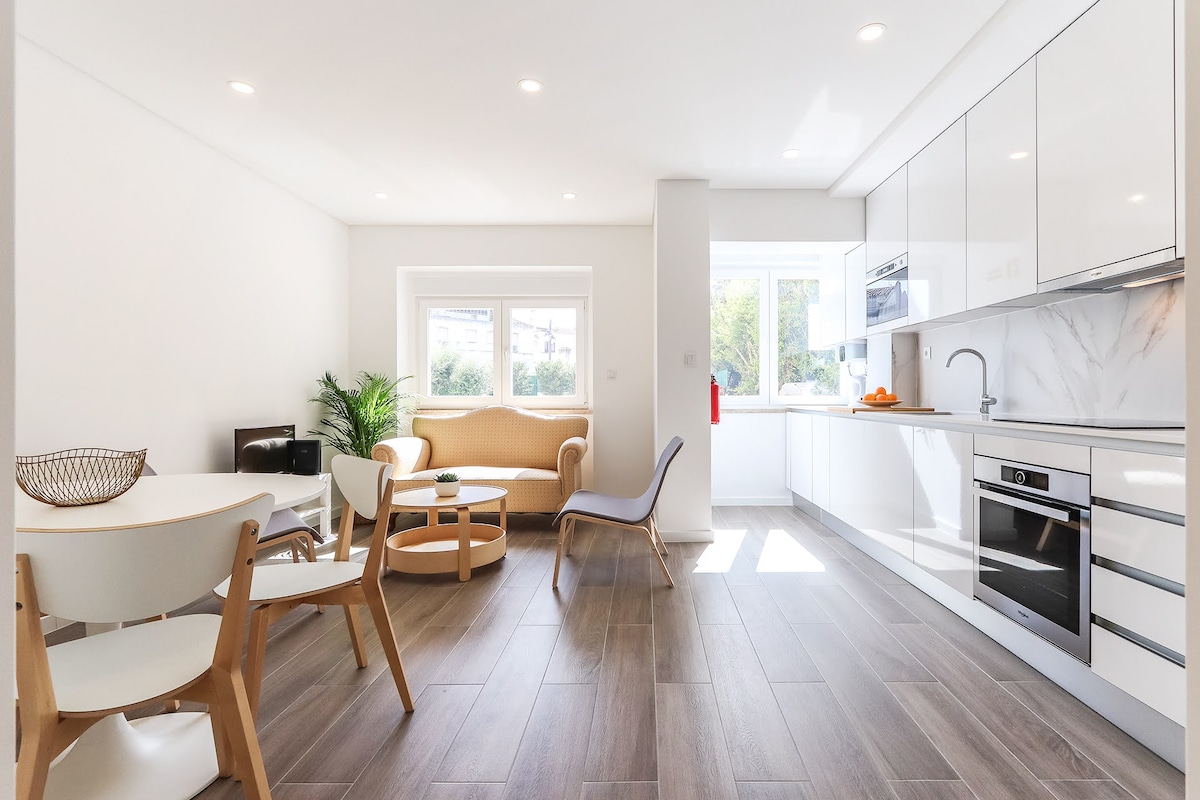 A modern kitchen and living area are presented, featuring sleek white cabinetry and appliances. A small dining table with light chairs is noticeable, accompanied by a cozy seating area with a beige couch. Natural light floods through the large window, enhancing the spacious feeling.