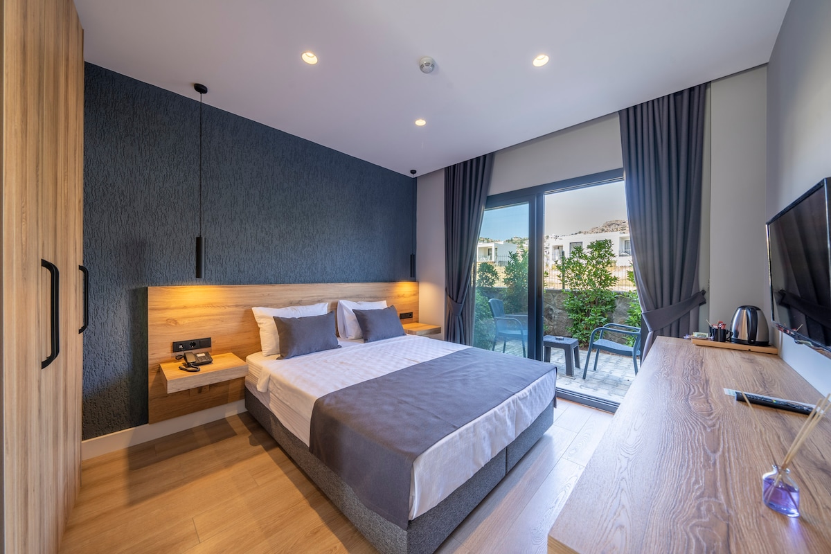 A modern bedroom is presented with a spacious bed adorned in neutral linens. Large glass doors lead to a private outdoor seating area, framed by elegant curtains. Wood accents are visible in the furniture, which includes a desk and a small coffee-making station.