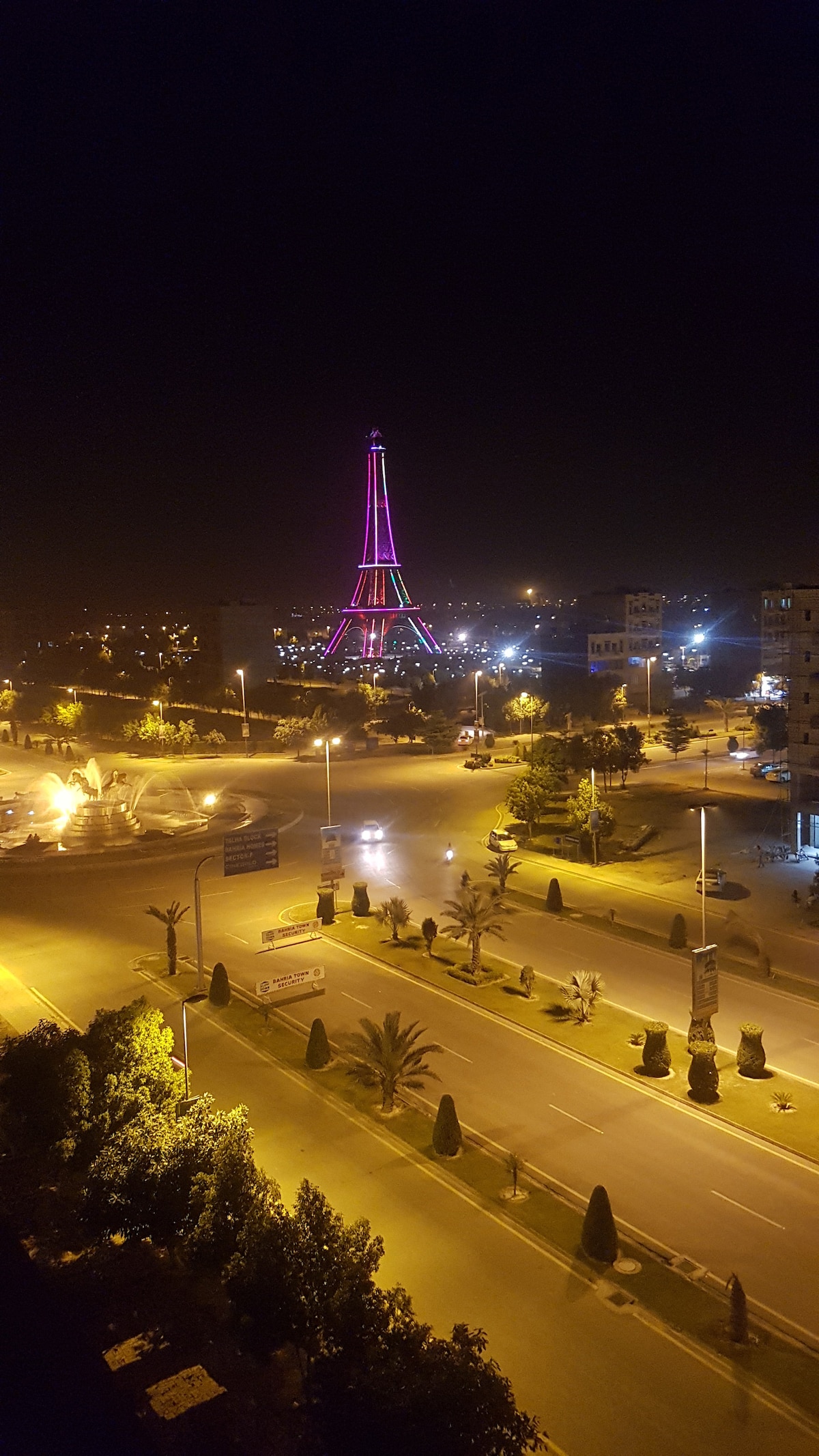 A vibrant view of a tower illuminated in pink hues against a night sky is presented. The surrounding streets, lined with streetlights and palm trees, add to the ambiance, while nearby buildings are softly lit, contributing to a serene urban landscape.