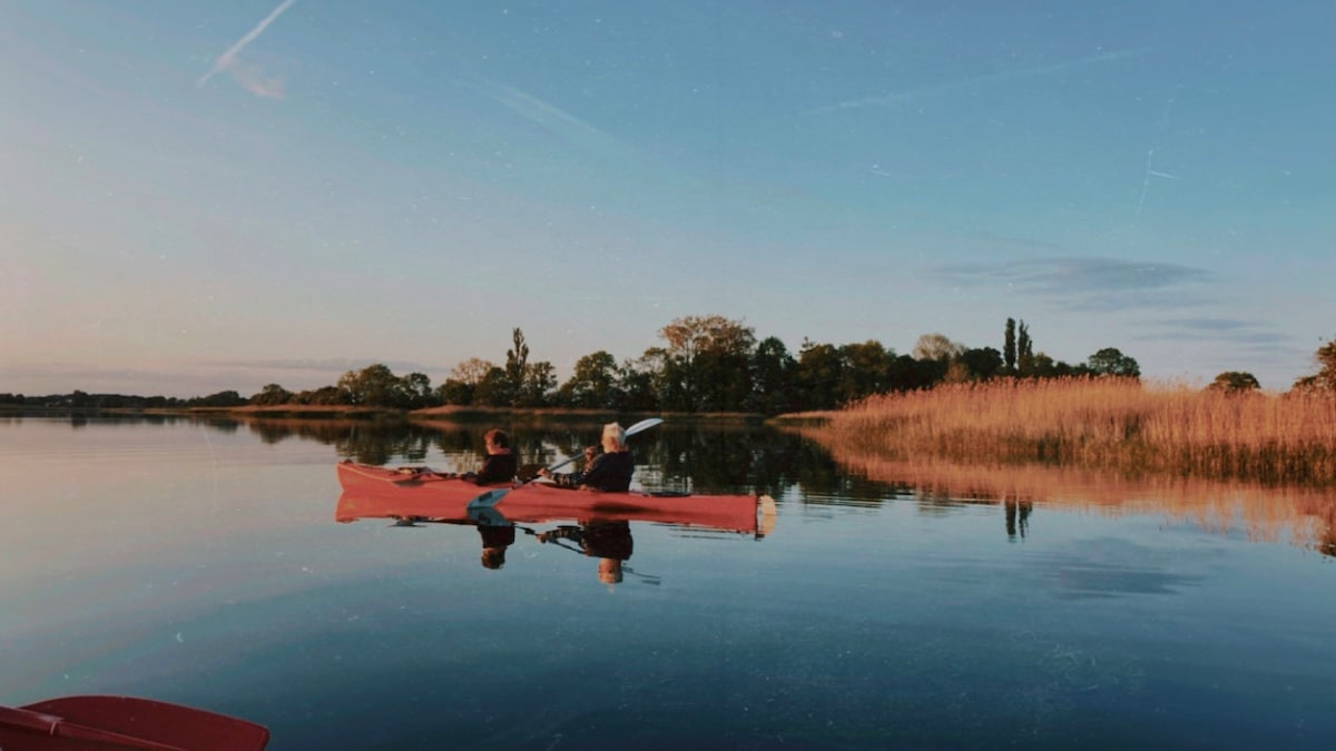 Two individuals are kayaking on calm waters during sunset, reflecting the warm hues of the sky. Lush greenery flanks the shores, while tall reeds create a natural border. The scene conveys a sense of tranquility and connection to nature.