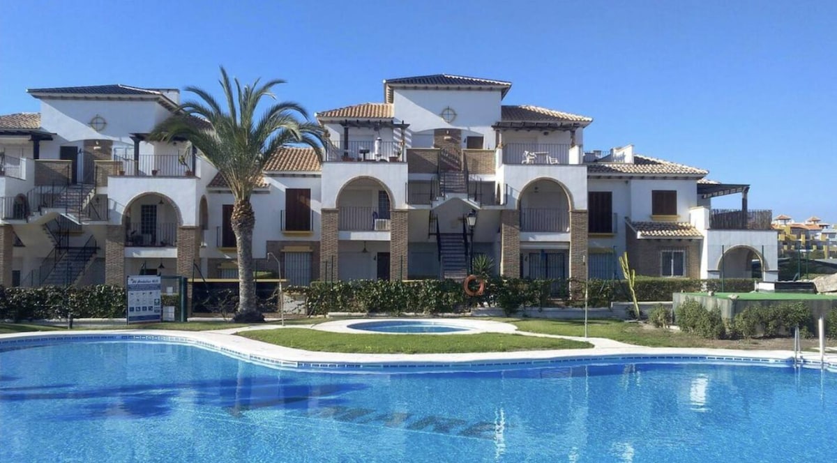 A private residential area features a modern building with multiple balconies and arches. An inviting outdoor pool is surrounded by landscaped gardens, with lounge chairs positioned nearby. Palm trees frame the scene under a clear blue sky.