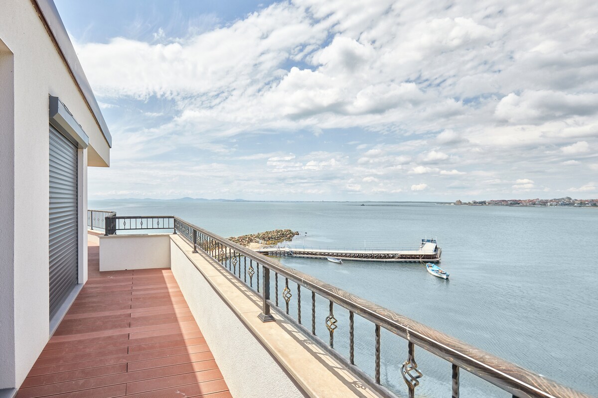 A balcony offers a clear view of the vast sea and a nearby boat pier. The railing and wooden flooring provide an elegant touch to the outdoor space. The sky is filled with clouds, enhancing the tranquil atmosphere.
