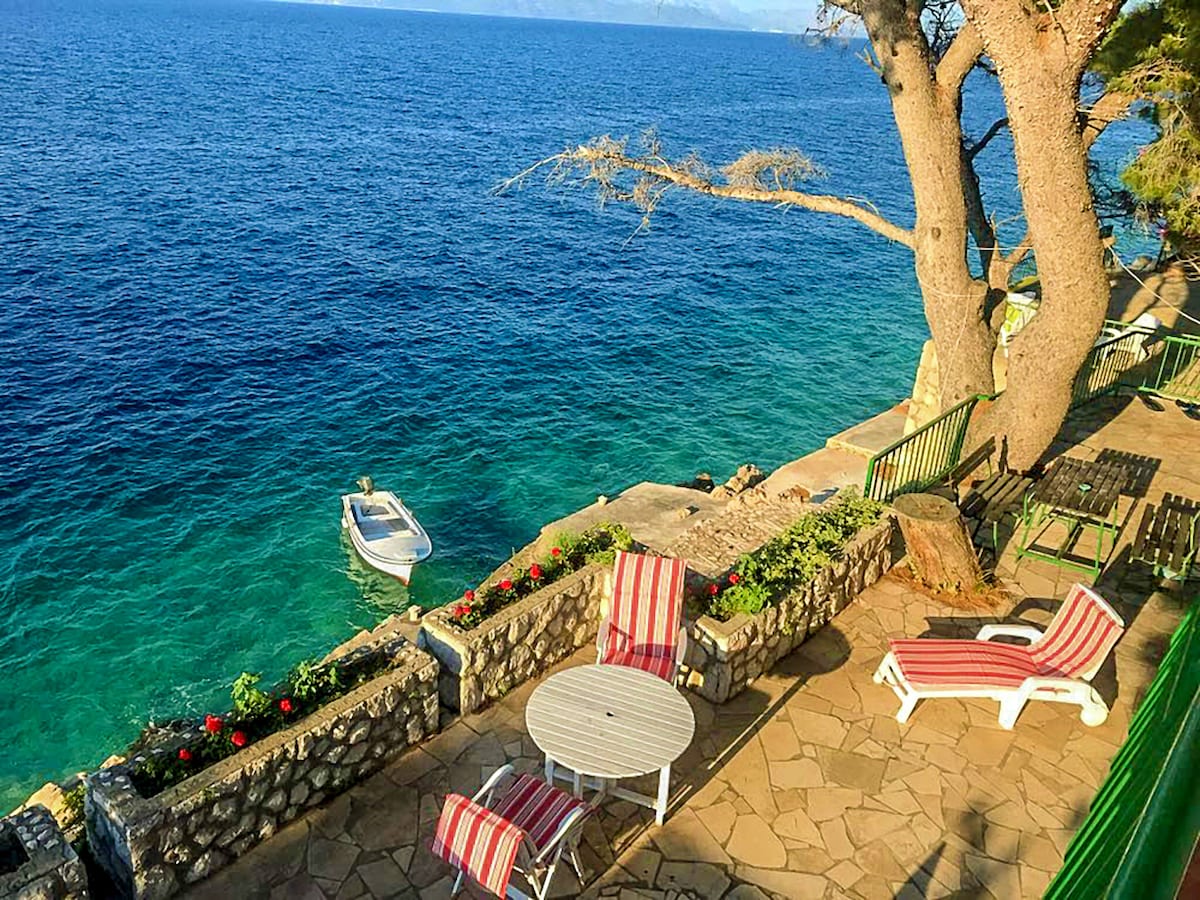 A seaside terrace is surrounded by stone walls and vibrant flowers, offering views of the clear blue Adriatic Sea. A small boat is moored nearby. The area features a circular table with striped chairs and reclining lounge chairs, providing a relaxing space for outdoor enjoyment.