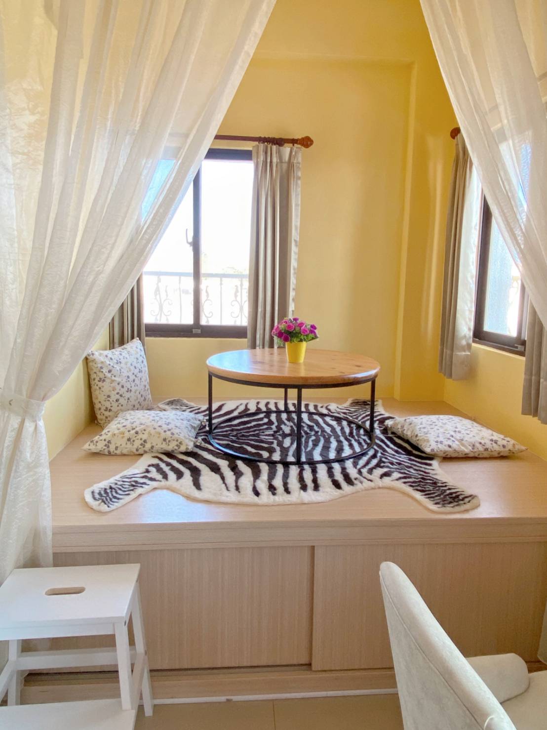 A cozy nook features a low wooden table surrounded by soft cushions on a raised platform. A zebra-patterned rug adds a unique touch. Natural light filters through the curtains, highlighting the bright yellow walls and a small vase of flowers on the table.