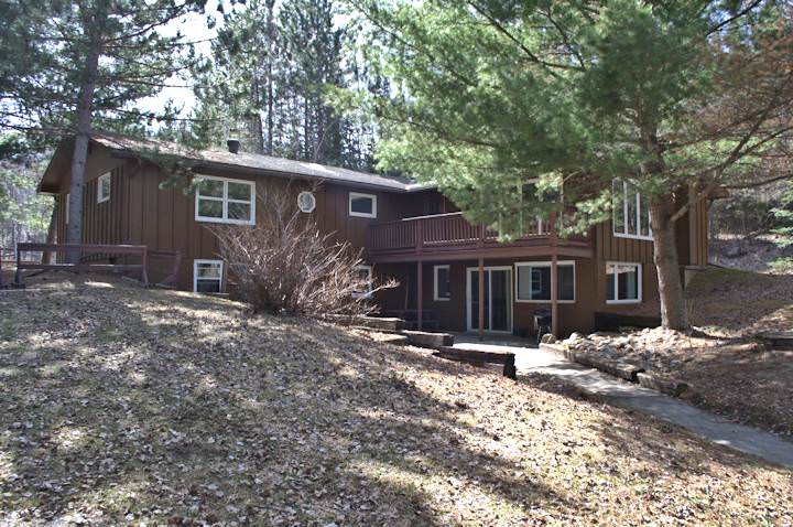 The exterior of the Retreat Cabin is surrounded by wooded areas, with large windows displaying a welcoming entrance. The cabin features a spacious deck and a sloping pathway leading through natural landscaping, emphasizing a serene and private setting.