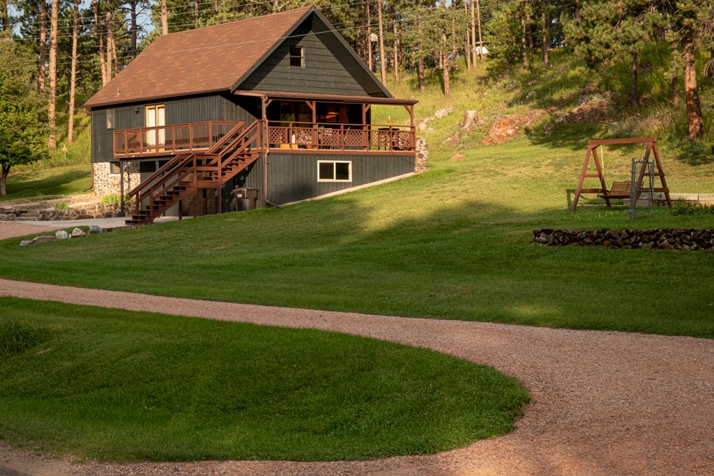 The cabin is nestled among trees, featuring a sloped lawn leading up to the dark exterior. A wooden deck is accessible by stairs at the front, and a swing set is positioned in the yard. Gravel pathways meander around the property.