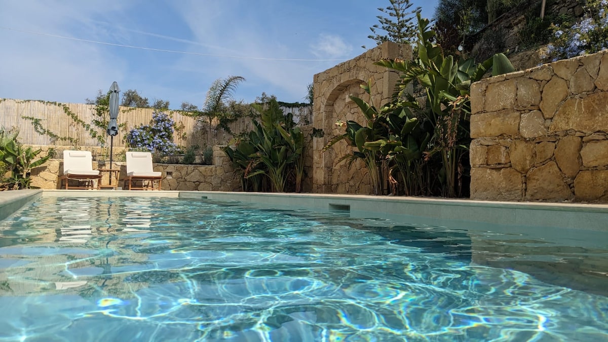 A serene swimming pool reflects blue skies and is framed by natural stone walls. Lush greenery surrounds the pool area, with two lounge chairs positioned nearby under the shade of an umbrella. The atmosphere is calm and inviting, perfect for relaxation.