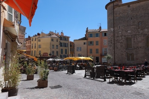 A lively outdoor plaza is shown, featuring multiple tables and chairs under large umbrellas. The surrounding buildings display a mix of colors, contributing to a vibrant atmosphere. Sunlight illuminates the scene, highlighting the cobblestone pathway and nearby greenery in decorative planters.