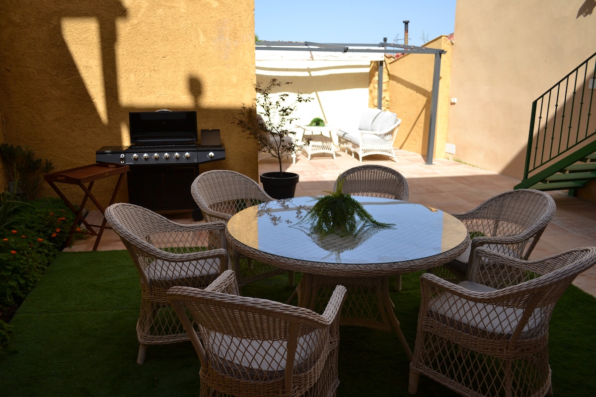 An outdoor seating area is showcased, featuring a round glass table surrounded by six woven chairs. A barbecue grill stands nearby, and a potted plant adds a touch of greenery. Natural light illuminates the space, creating an inviting atmosphere.