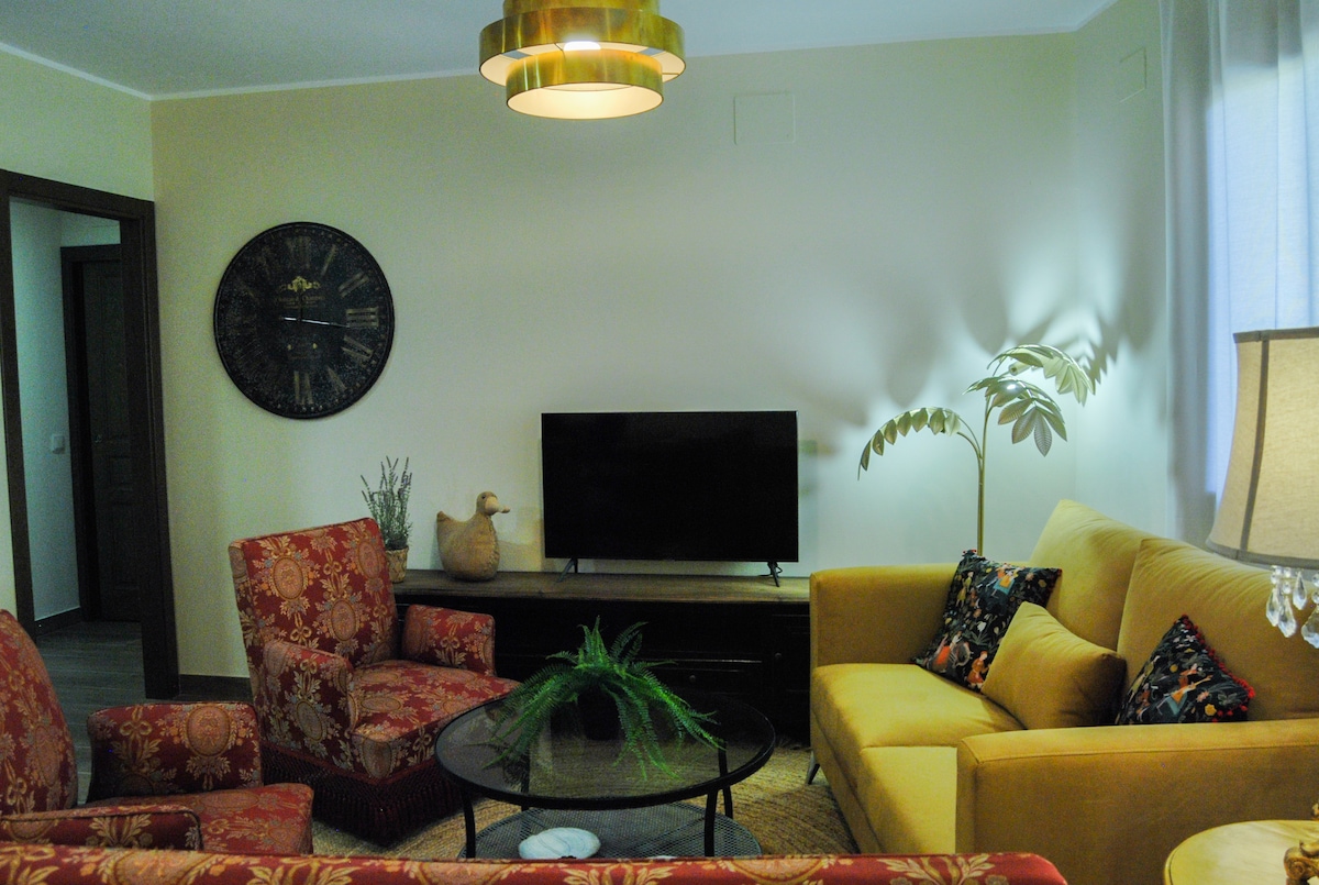 A bright living room features a golden light fixture hanging above a circular coffee table. Two patterned armchairs and a yellow sofa provide seating. A television is placed on a dark wooden console, complemented by a decorative plant and a wall clock.
