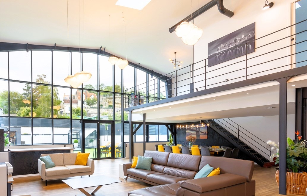 A spacious living area is highlighted by large windows that allow natural light to fill the room. Contemporary light fixtures hang from the high ceiling, while a comfortable sofa with bright cushions invites relaxation. The open layout extends to a dining area visible in the background.