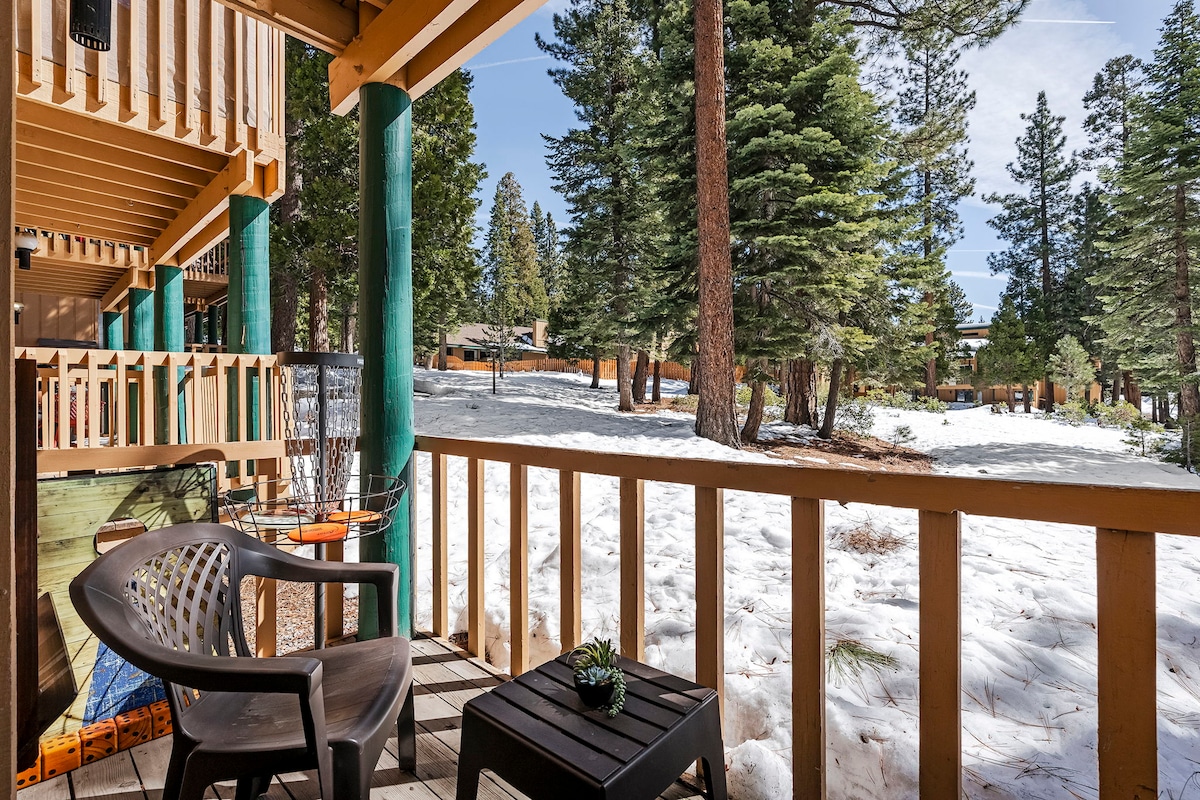 A charming outdoor balcony is surrounded by tall pine trees and a blanket of snow. A small table sits beside a black chair, with a decorative plant adding a touch of green. The serene winter landscape offers a peaceful retreat.