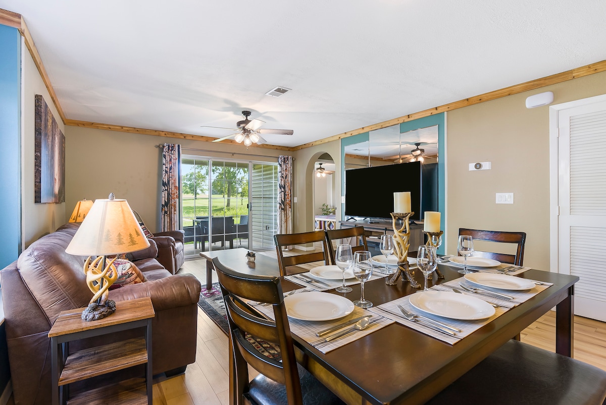 A dining area is set with elegant dinnerware and glassware on a dark wooden table. The space features a comfortable leather sofa, and large windows provide a view of the outdoors. Ceiling fans and decorative accents contribute to the inviting atmosphere.