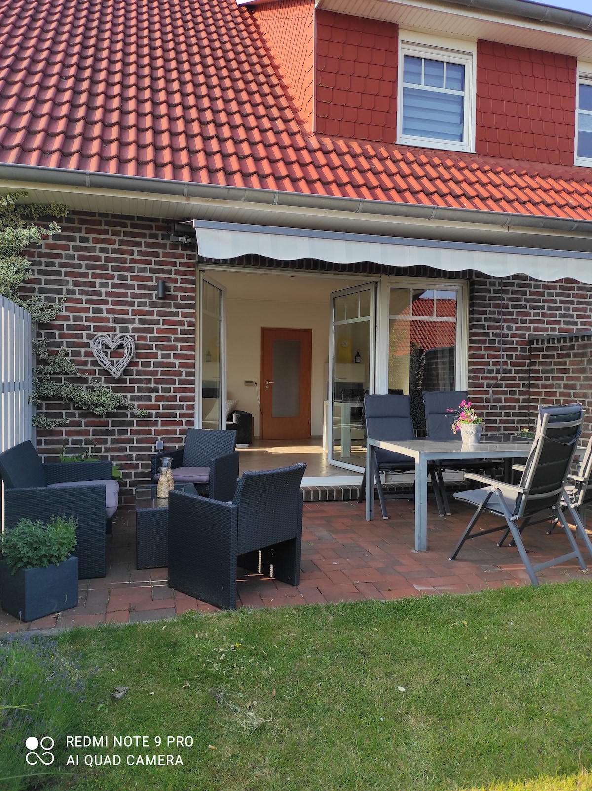 A patio area is featured with black wicker seating and a table, surrounded by a grassy lawn. A partial view of a brick house is visible in the background, with large windows allowing natural light into the interior. An awning provides shade, enhancing outdoor comfort.