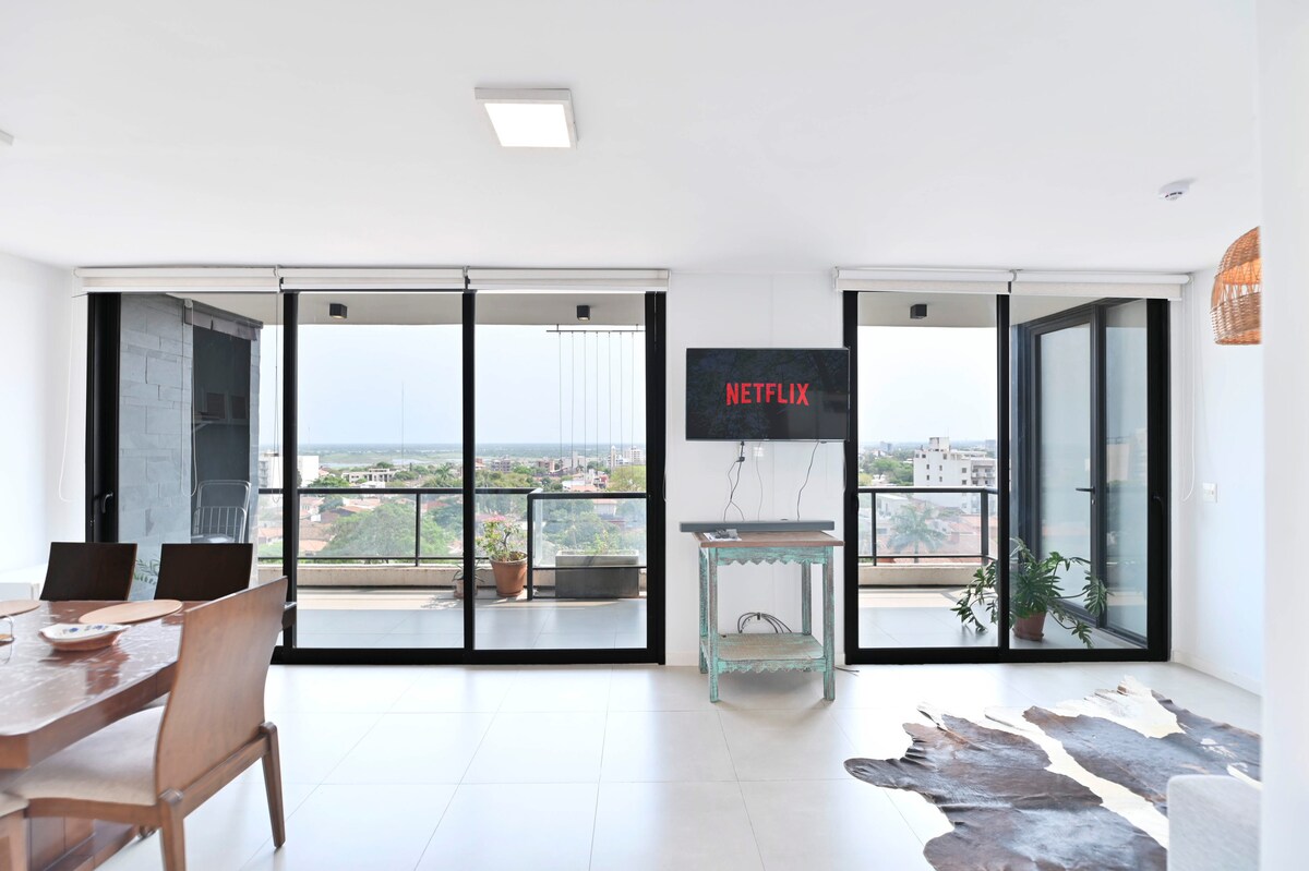 A modern living area showcases large glass doors that offer expansive views of the surrounding area. A wooden dining table is set for several guests. A television mounted on the wall displays the Netflix logo. Natural light fills the space, highlighting the sleek design.