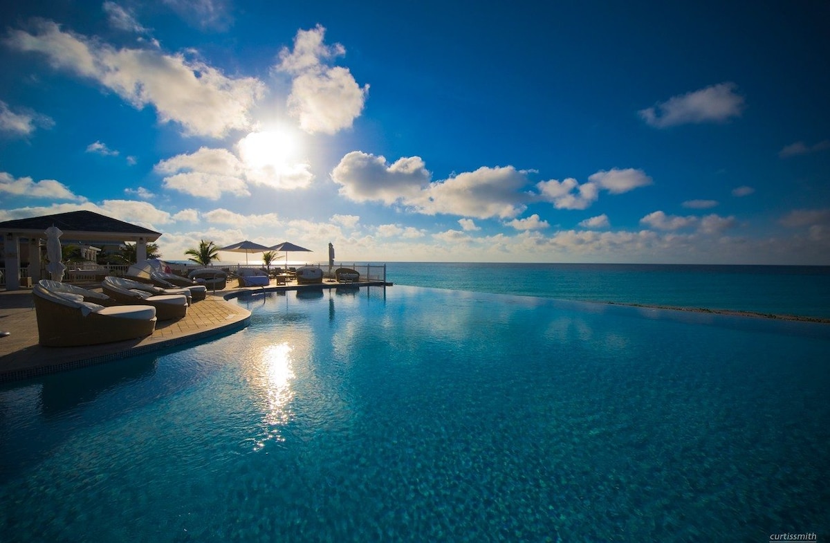 An infinity pool reflects the clear blue sky, with the sun positioned on the horizon. Lounge chairs are arranged around the pool, providing a space for relaxation. Umbrellas offer shade near the water's edge, enhancing the tranquil atmosphere of the scene.