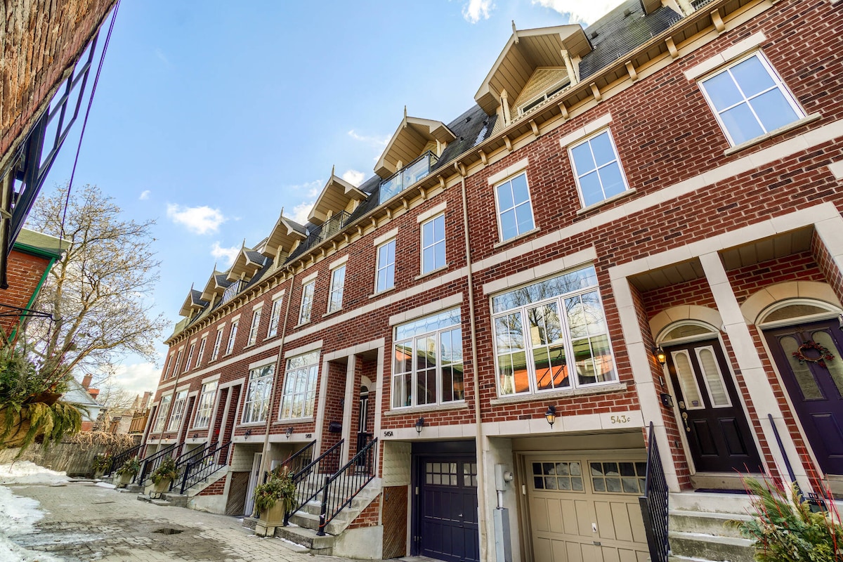 The multi-story townhouse, characterized by red brick exteriors and large windows, showcases a row of three distinct units. A welcoming pathway leads to the entrance, flanked by planters. The clear blue sky overhead complements the modern architectural design.