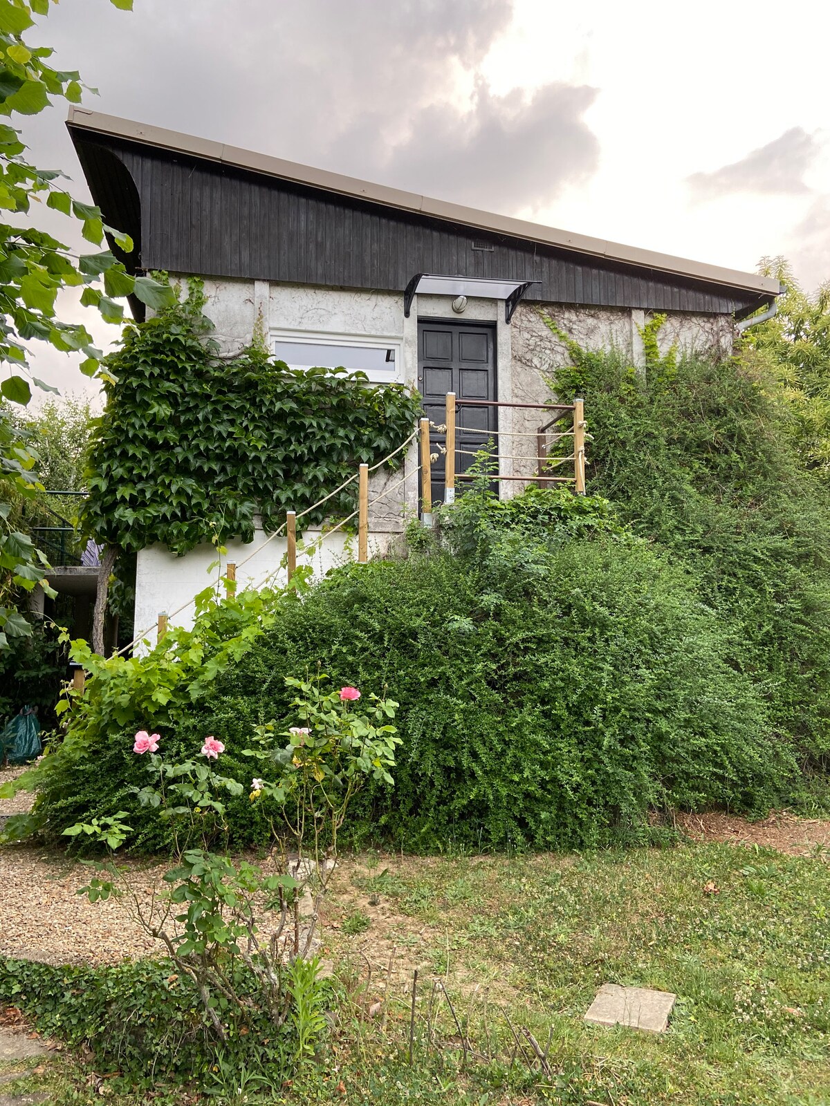 A charming two-story house, surrounded by lush greenery, features a dark wooden roof and a front staircase leading to the entrance. Vibrant pink flowers are visible in the garden, enhancing the serene outdoor setting.