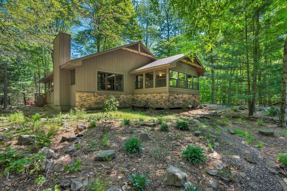 A family cabin is nestled among lush greenery, with large windows allowing natural light to fill the interior. The exterior features a combination of stone and wood finishes, and the surrounding area includes landscaped rocks and a variety of plants, enhancing the tranquil forest setting.