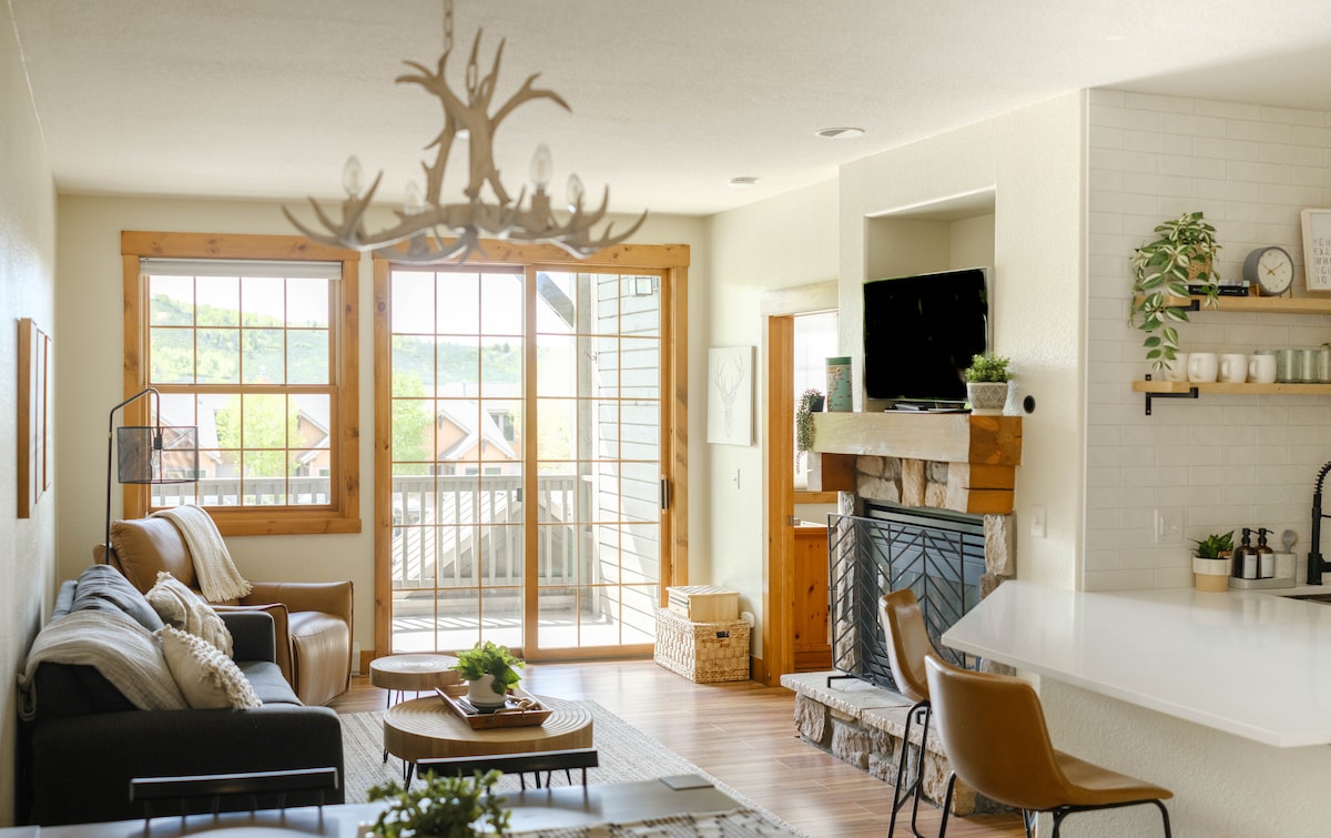 A bright living area is presented with two comfortable sofas, arranged around a central coffee table. Large windows allow natural light to illuminate the space, revealing views of the outdoors. A modern fireplace and an open kitchen area enhance the inviting atmosphere.