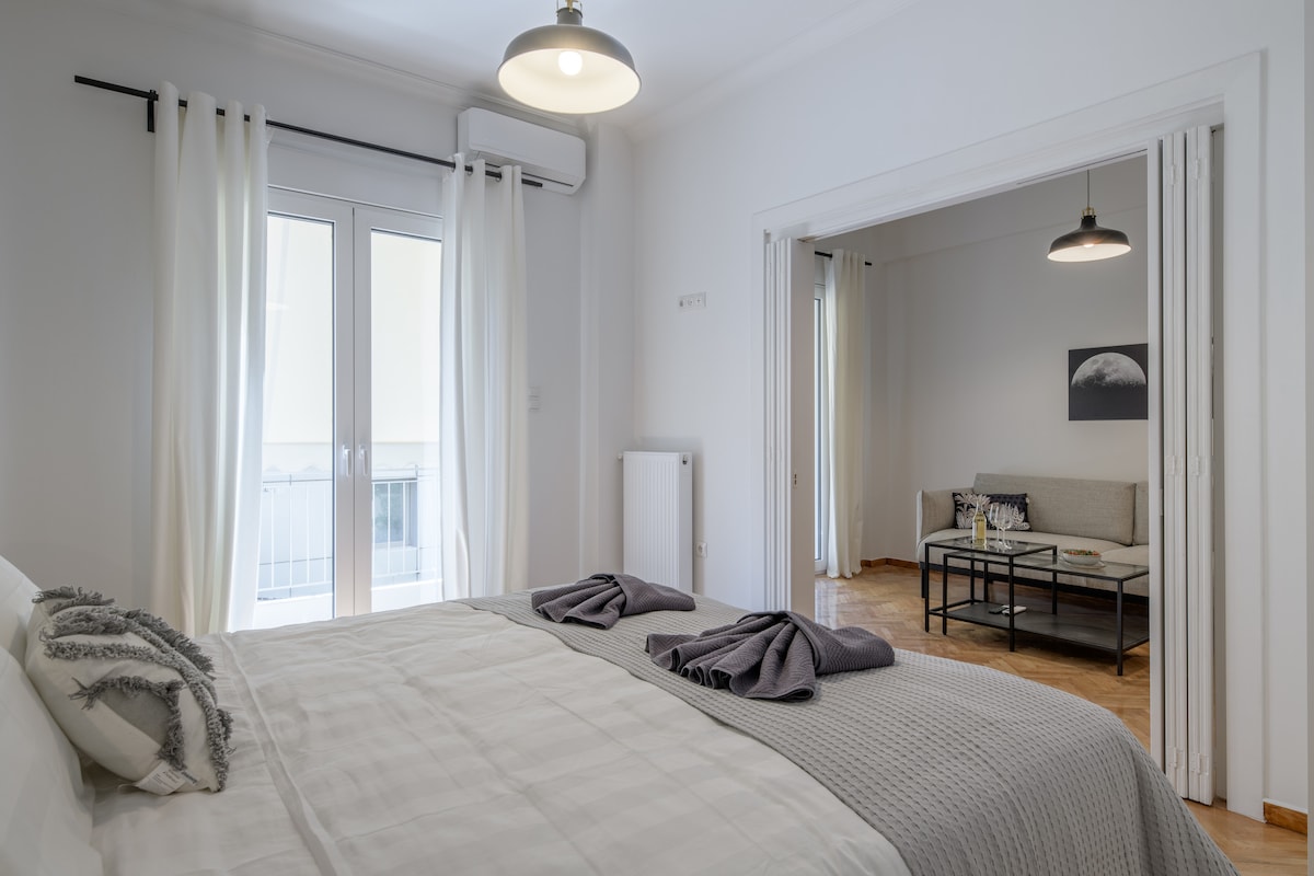 A spacious bedroom features a large bed with neatly arranged towels. Light-colored curtains frame a set of glass doors leading to a balcony. The adjoining living area is visible, containing a couch and a coffee table, enhancing the apartment's open layout.