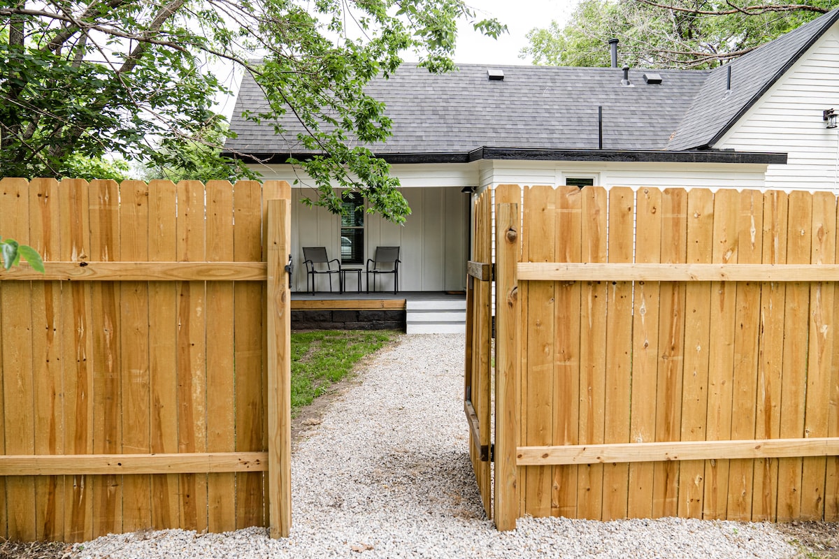 A wooden fence with a gate opens to a gravel pathway leading to the house. The exterior features a porch with seating, surrounded by greenery and trees, providing a welcoming entryway to the property.