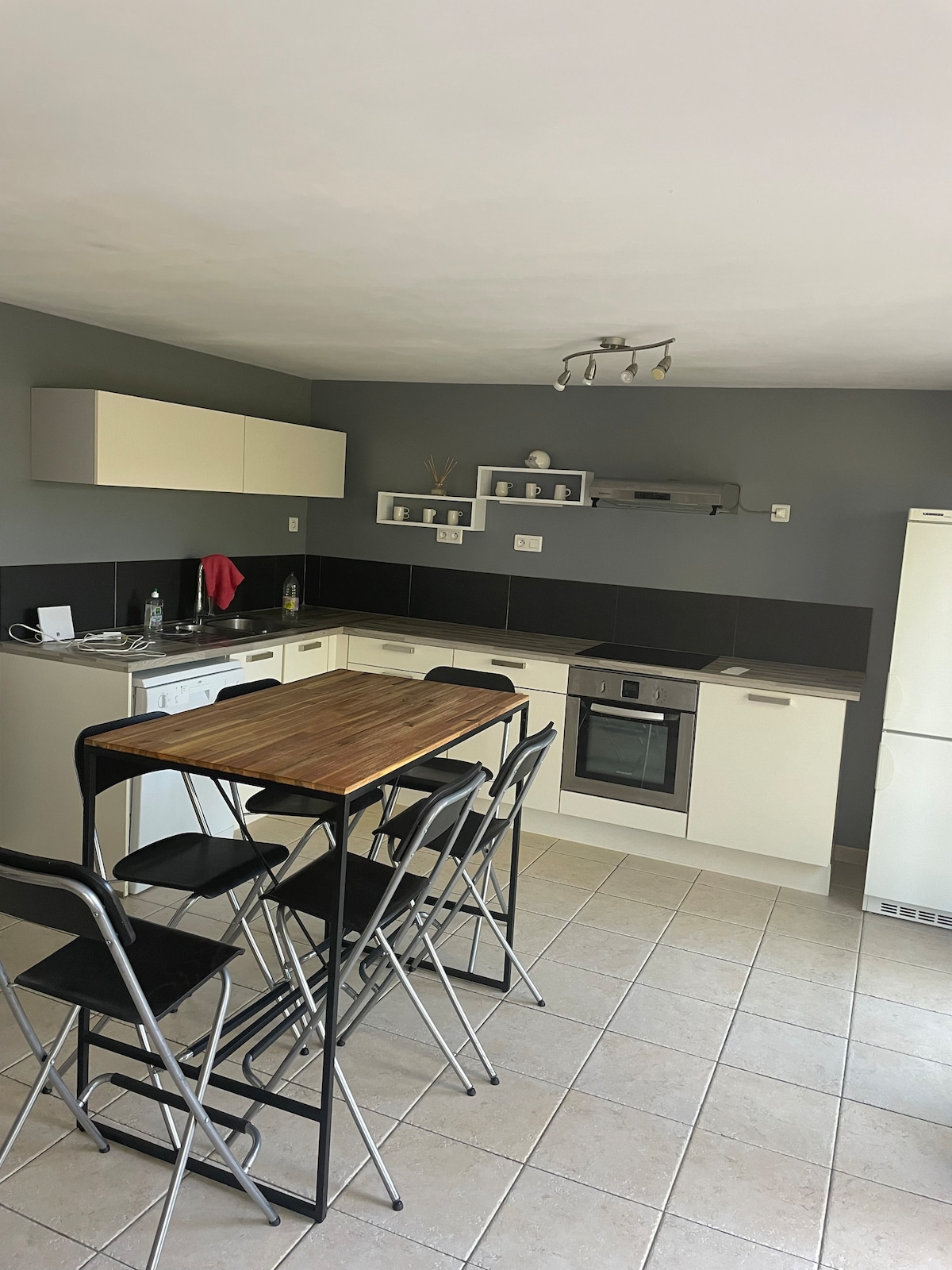 The kitchen showcases a modern design with light cabinetry and dark countertops. A compact dining table is accompanied by several black stools. Open shelves hold decorative items, while a spacious refrigerator is positioned nearby, contributing to a functional and inviting space.