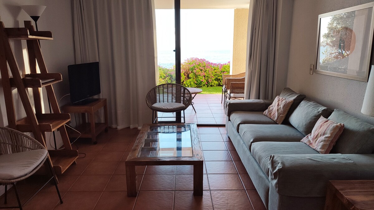 A living area is presented with a comfortable sofa and two chairs. A glass coffee table sits in the center. Large sliding doors open to a terrace with views of the sea, framed by sheer white curtains, allowing natural light to fill the space.