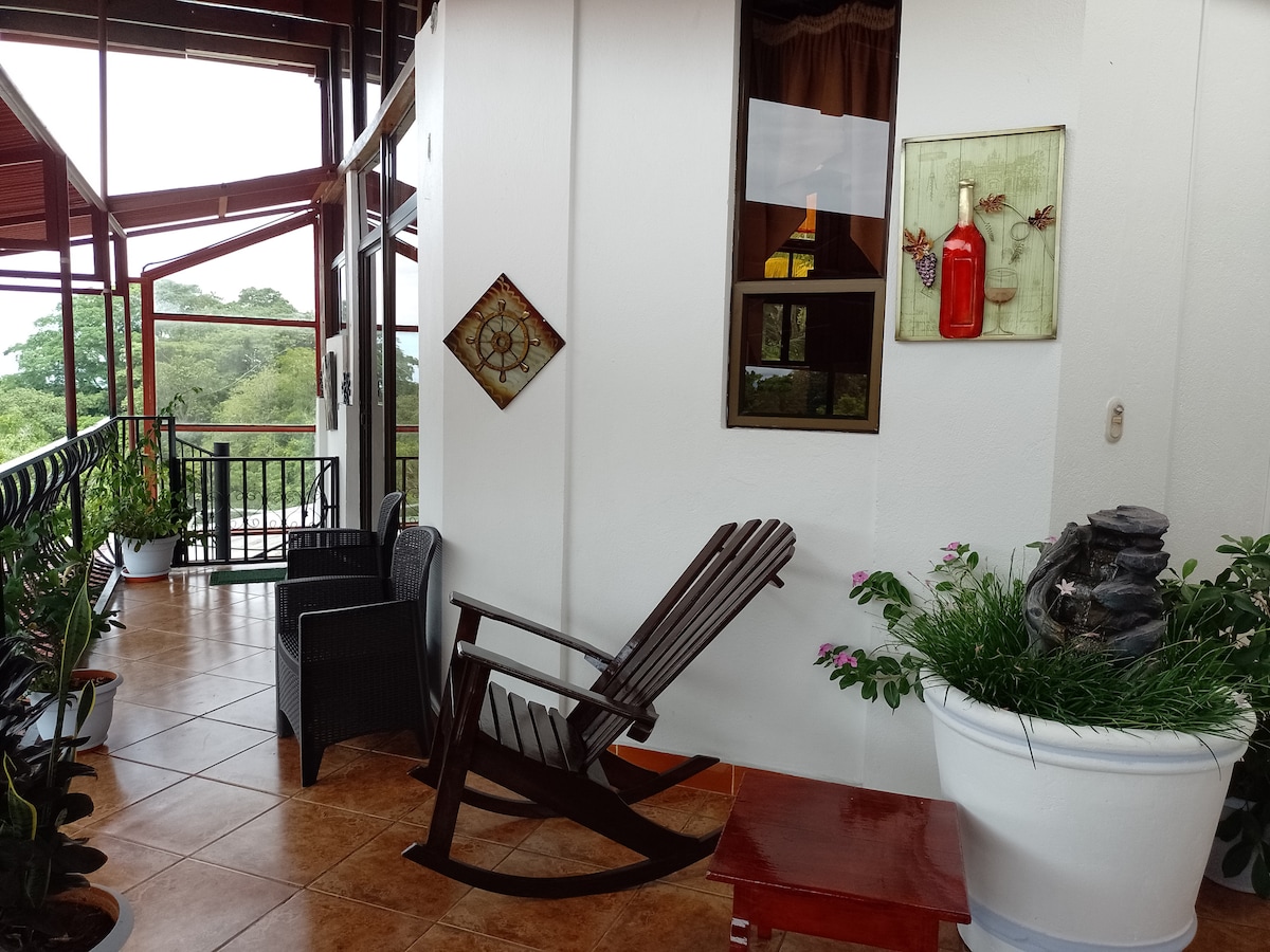 A spacious balcony is shown, featuring a wooden rocking chair and two wicker chairs, inviting relaxation. Decorative items, including a small wall art piece, add character. Lush greenery surrounds the area, and a serene stone feature can be seen nearby, enhancing the tranquil setting.