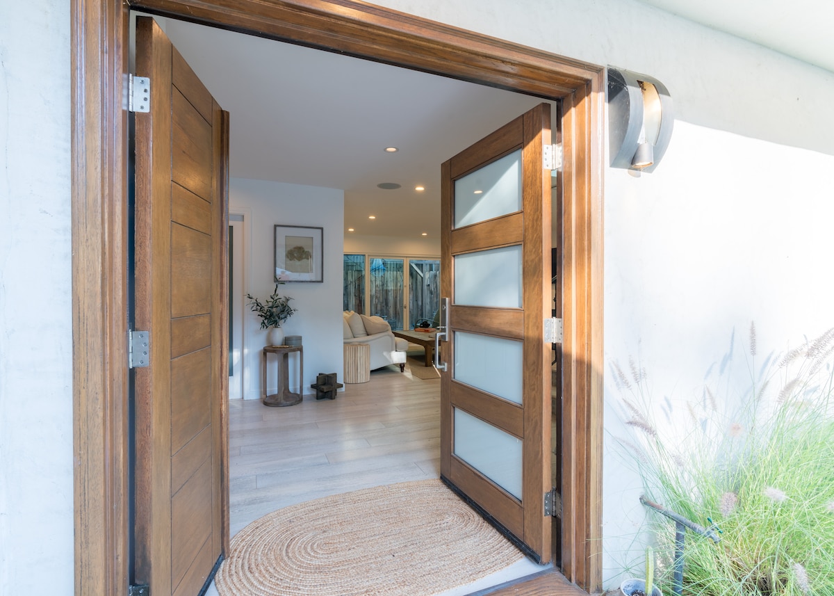 The entrance showcases a set of elegant wooden doors, opened wide to reveal a bright living area. Soft light illuminates the space, highlighting a cozy seating arrangement and natural decor elements. A round woven mat lies at the threshold, adding texture to the welcoming atmosphere.