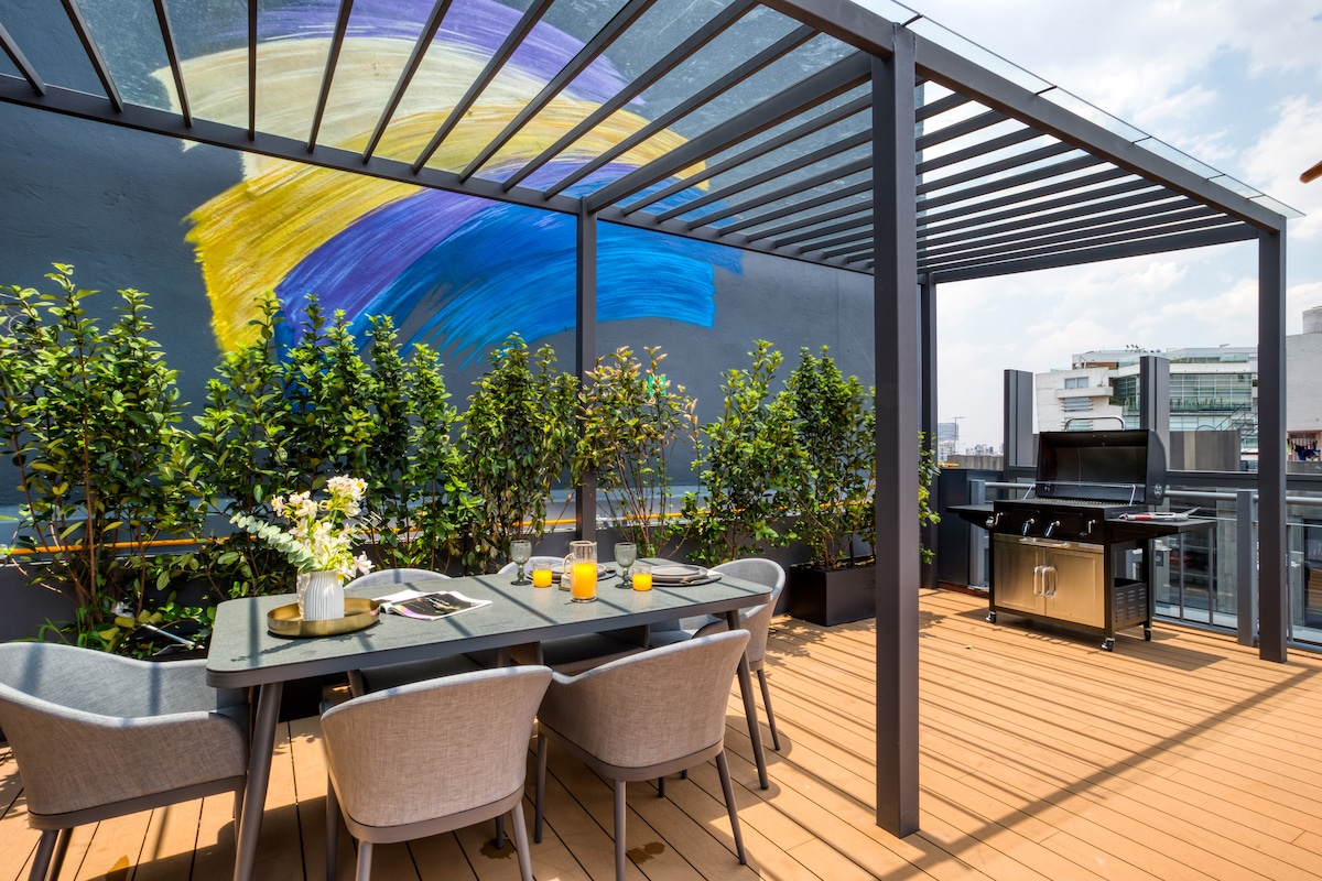 A private rooftop area features a dining table with comfortable chairs, surrounded by greenery. An outdoor barbecue grill is present alongside a striking mural in vibrant colors. The wooden deck offers ample space for relaxation and outdoor dining.