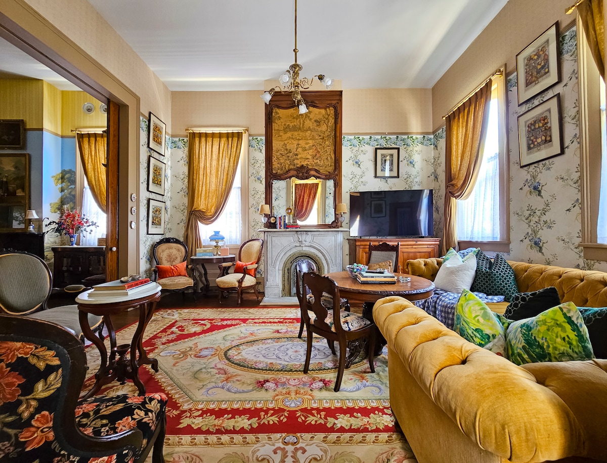 A spacious parlor is furnished with vintage seating and a large area rug featuring intricate patterns. The walls are adorned with framed artwork, and large windows are dressed with elegant drapes, allowing natural light to filter softly into the room. A marble fireplace serves as a focal point.