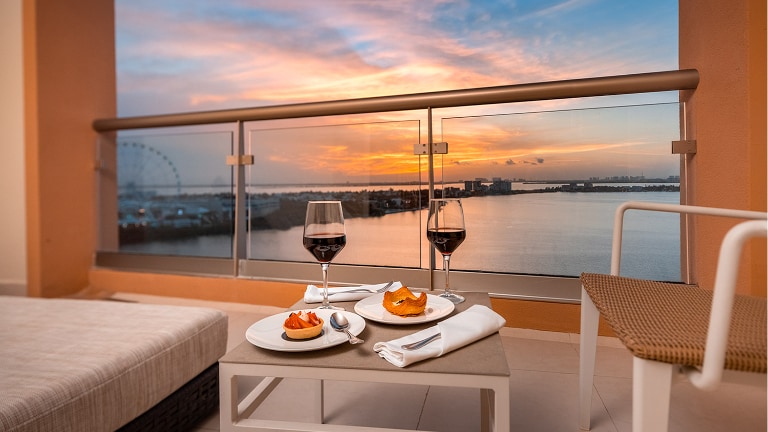 A serene balcony view captures a sunset over the water, featuring a small table set with two glasses of red wine and plates of food. The scene is complemented by soft seating and a railing that frames the expansive landscape.