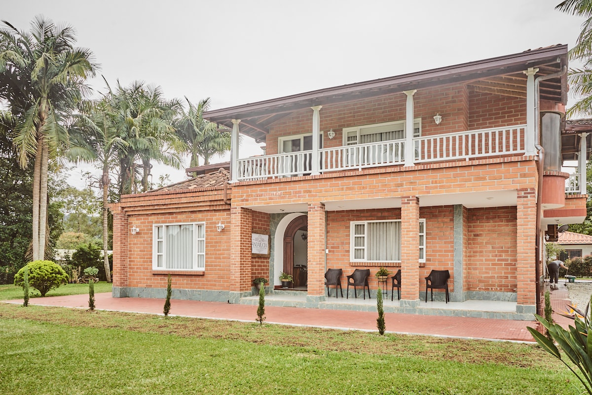 A two-story brick house is surrounded by lush greenery and palm trees. A spacious outdoor area features well-maintained grass and neat hedges. The front porch is furnished with several chairs, inviting relaxation. Large windows provide ample natural light to the interior.