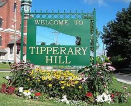 A green welcome sign for Tipperary Hill is showcased, featuring white lettering on a vibrant background. Colorful flowers in red, yellow, and purple bloom around the base of the sign, complemented by lush green grass. The sky above is bright and clear.