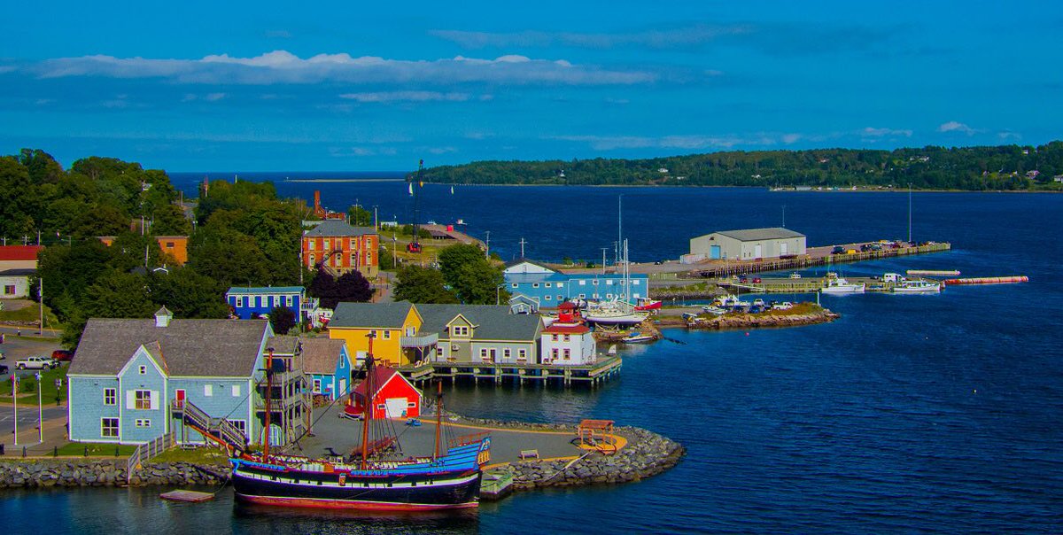 A diverse collection of colorful buildings lines the waterfront, with a mix of boats docked in the calm blue waters. The scene is framed by green hills in the background, and a clear blue sky stretches overhead, creating a serene coastal atmosphere.